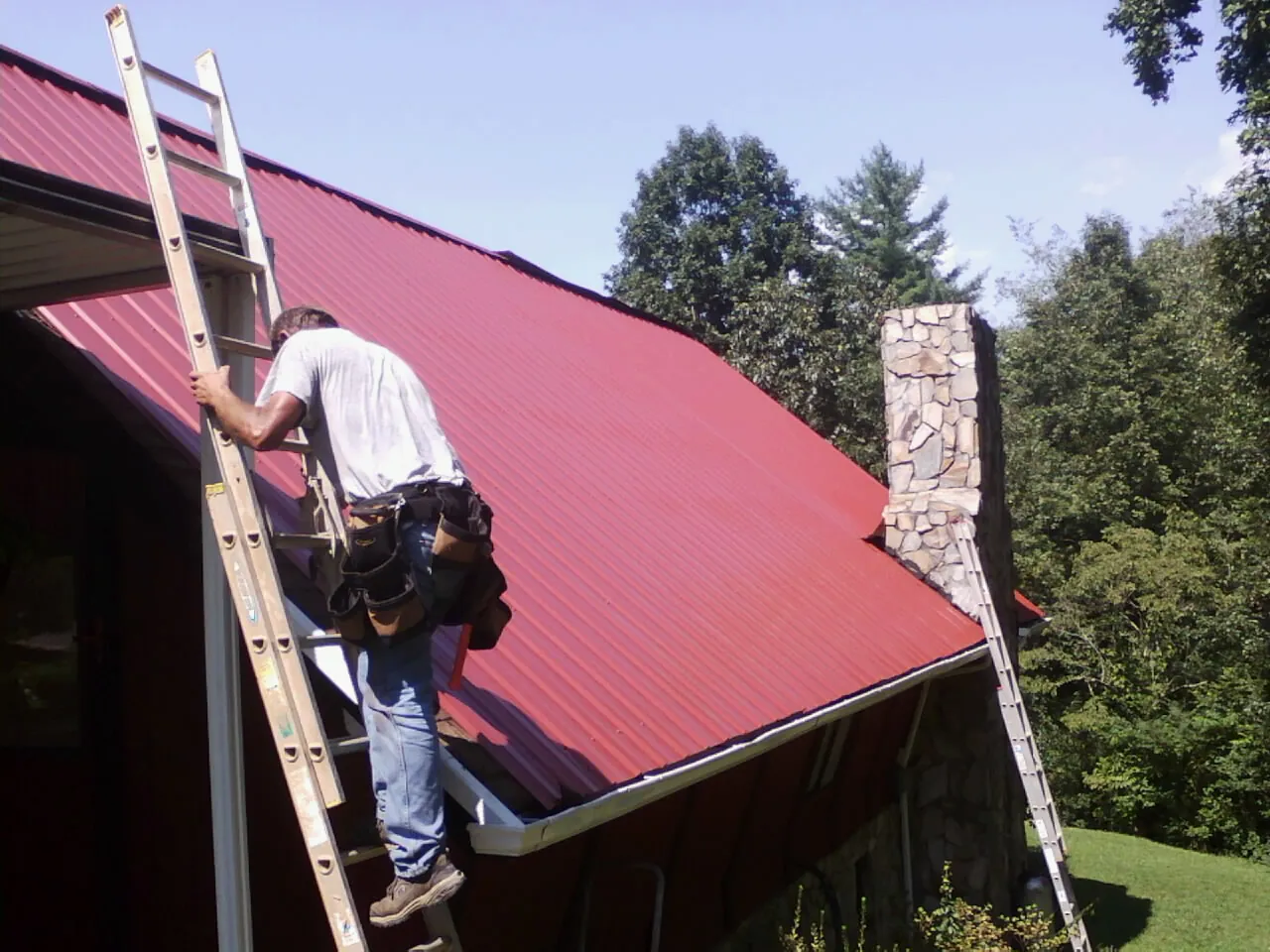 Roofer on a ladder beside a red metal roof and stone chimney — Soffit & Fascia Repair by Roofing Brothers in Saint Clair